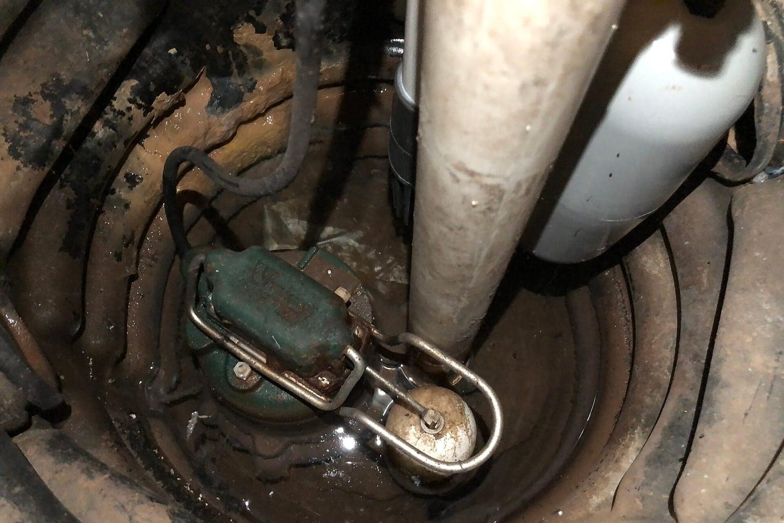 Submersible sump pump with a float switch sitting in a basement sump basin beside a white PVC discharge pipe